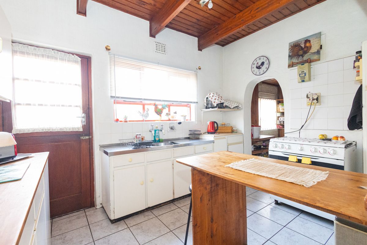 A dated kitchen featuring exposed timber ceiling beams, tiled flooring, a stainless steel sink, and a freestanding stove, with an archway leading to a scullery.