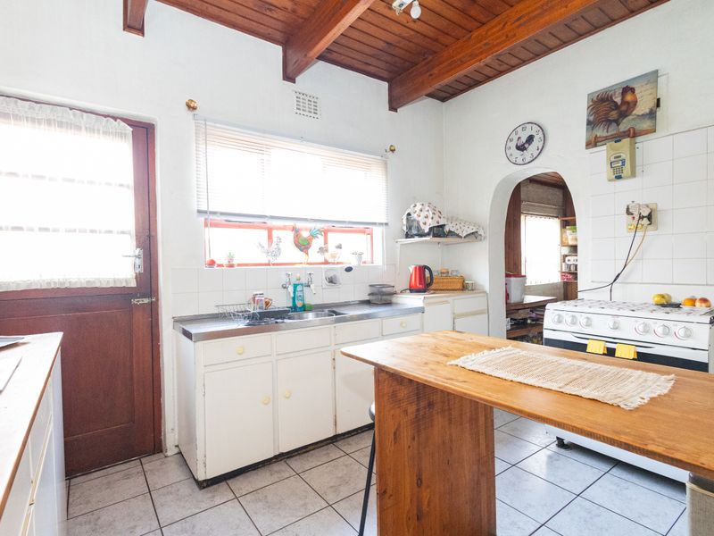 A dated kitchen featuring exposed timber ceiling beams, tiled flooring, a stainless steel sink, and a freestanding stove, with an archway leading to a scullery.