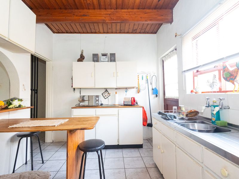 A functional kitchen featuring white cabinetry, a stainless steel sink, tiled flooring, and exposed timber ceiling beams, offering potential for modernization.