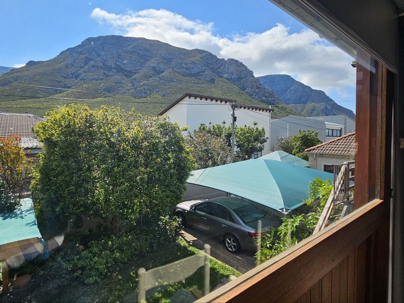 A scenic view from an interior window overlooking a residential garden, driveway with shade sail, and prominent mountain backdrop.