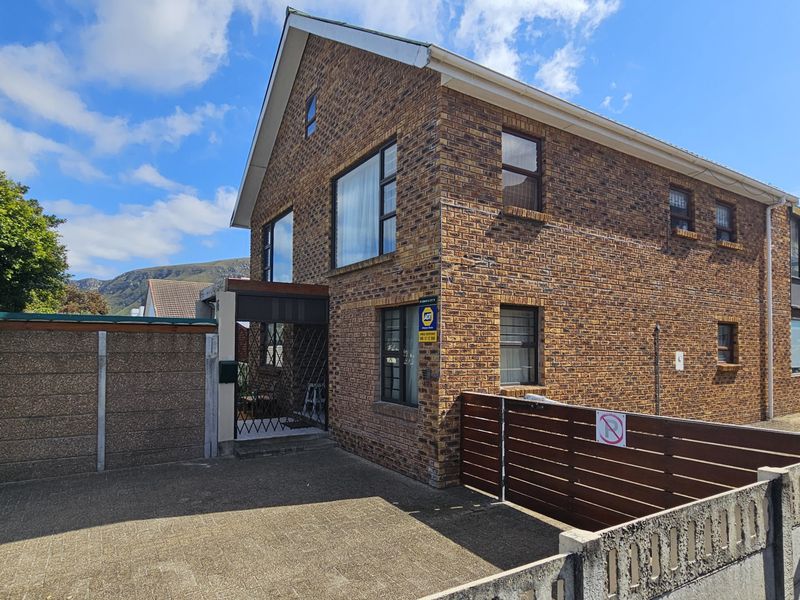 A double-storey face brick house featuring a paved driveway, secure perimeter walling, and aluminium window frames, set against a mountain backdrop.