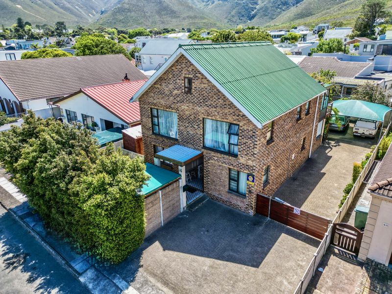 Aerial view of a double-story brick house featuring a green corrugated metal roof, paved driveway, and secure perimeter fencing in a residential neighborhood.