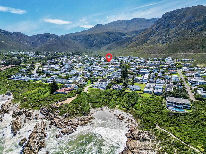 An aerial view of a coastal residential neighborhood nestled between the ocean and mountain range, with a location marker indicating the property.