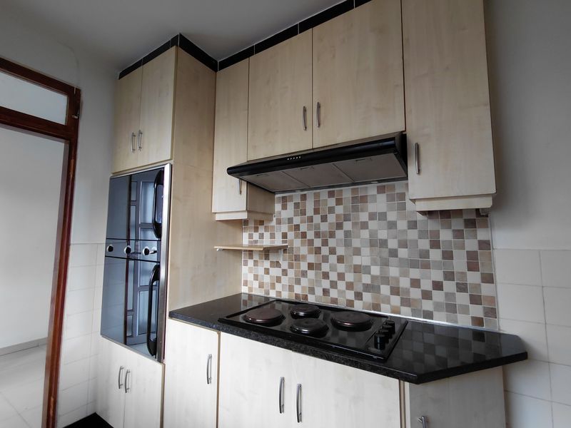 A functional kitchen featuring light wood cabinetry, dark laminate countertops, a built-in oven, electric hob, and a decorative tiled backsplash.