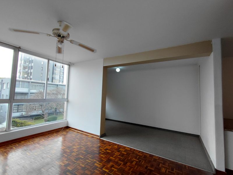 An empty living area featuring polished parquet flooring, a ceiling fan, and large windows providing natural light and an urban view.