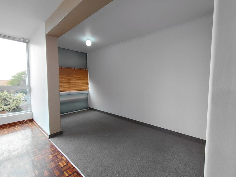 An empty living area featuring a combination of parquet and carpeted flooring, large windows with venetian blinds, and neutral wall finishes.