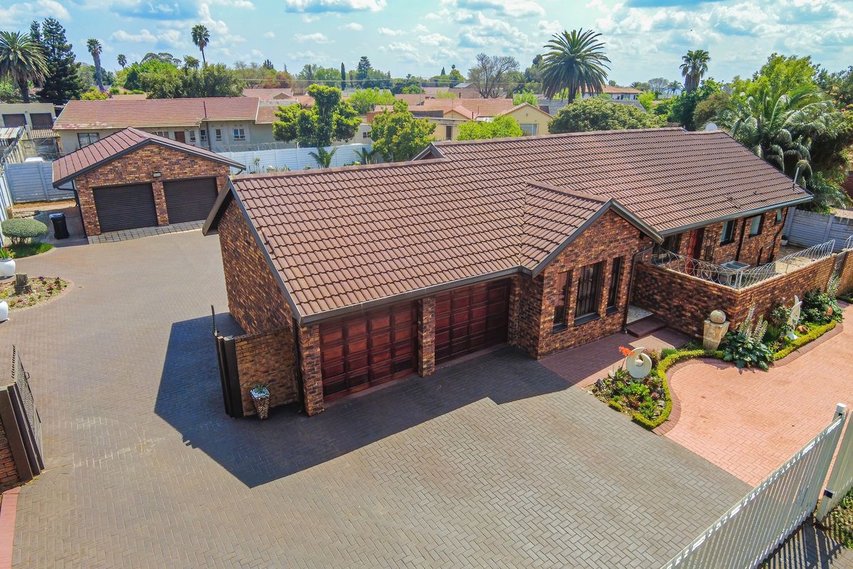 Aerial view of a well-maintained brick house featuring a tiled roof, double garage, extensive paved driveway, and established garden areas.