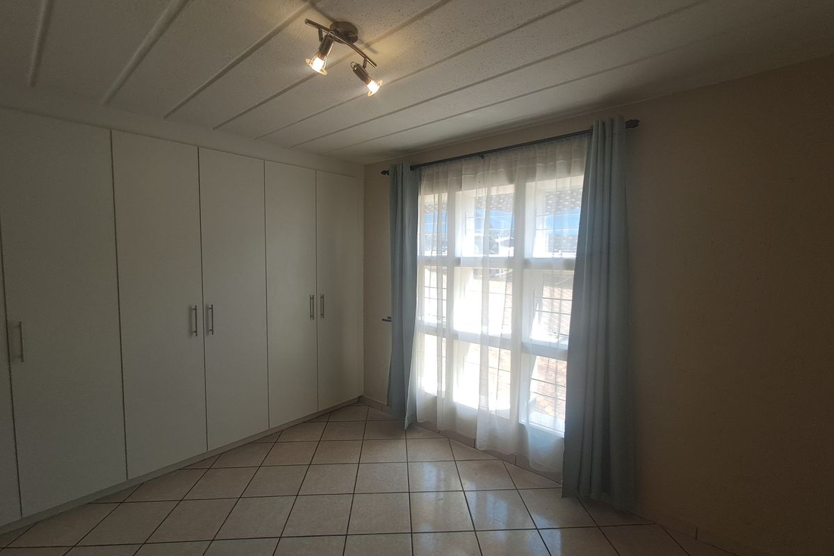 A bright, empty bedroom featuring tiled flooring, extensive built-in cupboard storage, and a window fitted with security bars.