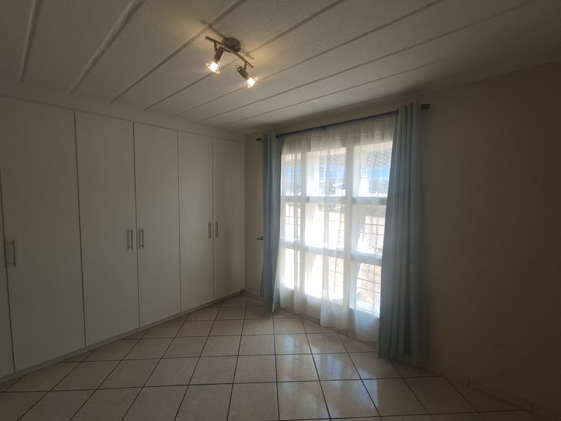 A bright, empty bedroom featuring tiled flooring, extensive built-in cupboard storage, and a window fitted with security bars.