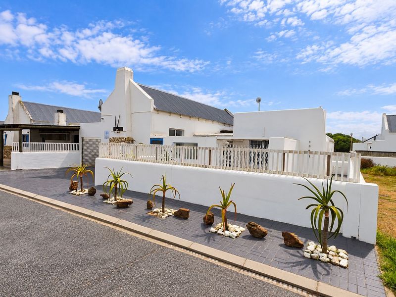 A well-maintained white cottage-style house featuring a corrugated metal roof, paved driveway, and a secure boundary wall with picket fencing.
