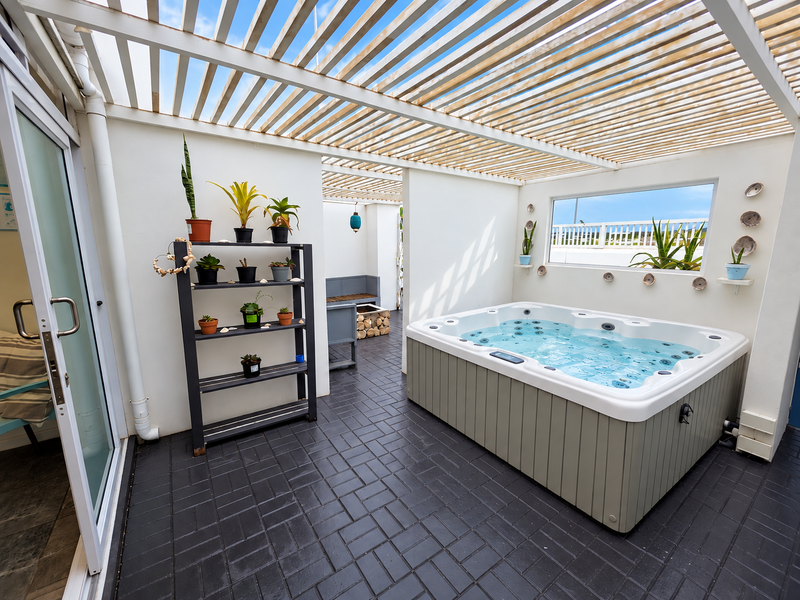A well-maintained covered patio featuring a built-in hot tub, slatted timber pergola, and dark paved flooring, offering a private outdoor relaxation space.