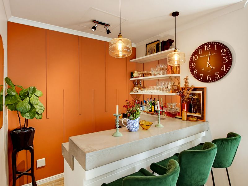 A stylish home bar area featuring a concrete countertop, built-in cabinetry with a vibrant orange accent wall, and modern pendant lighting.