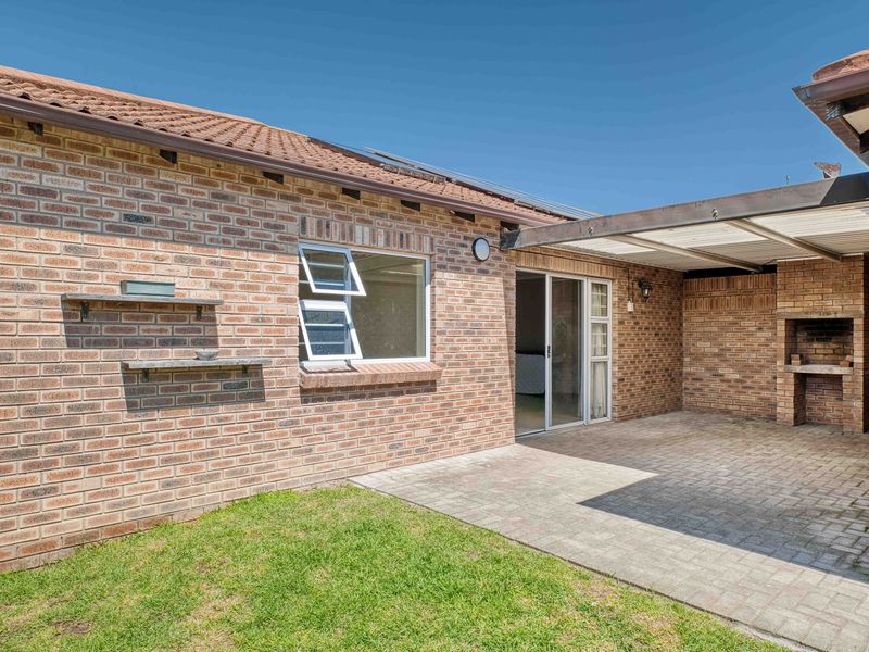 A well-maintained rear exterior featuring a paved patio area with a built-in braai, sliding door access, and a neat lawn.