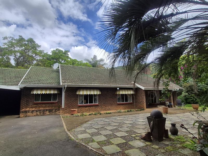 A single-story brick house featuring a tiled roof, attached carport, and a paved front courtyard area surrounded by established garden greenery.