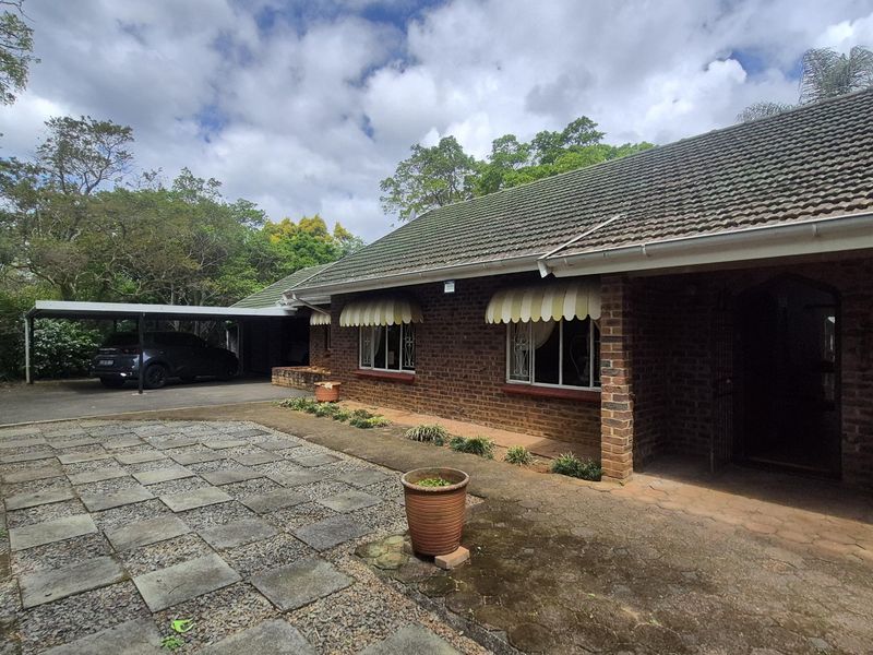 Front elevation of a brick house featuring a tiled roof, window awnings, security gates, and an attached carport with a paved driveway area.