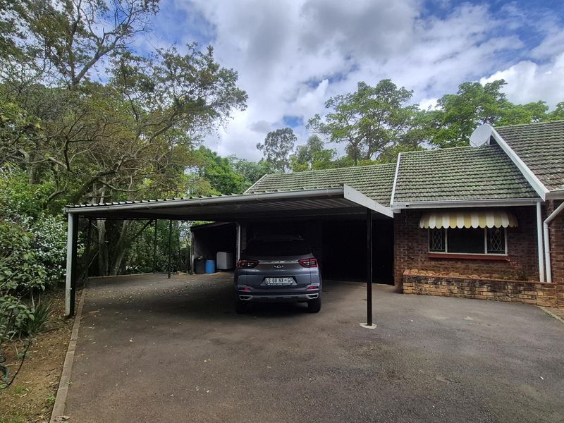 Front exterior view of the property featuring a covered carport, brick facade, and asphalt driveway surrounded by mature trees.