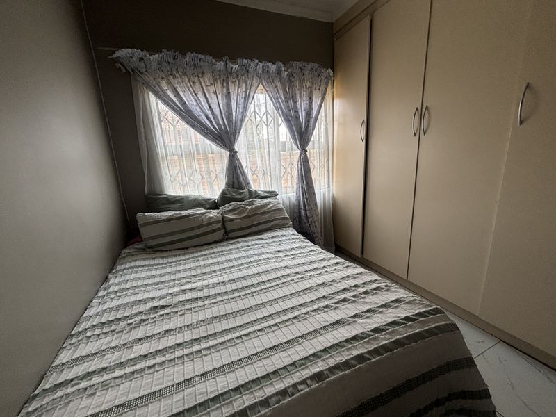 A compact bedroom featuring built-in cupboards, tiled flooring, and a window fitted with a security gate.