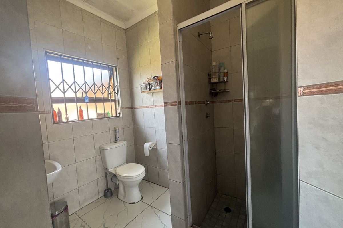 A functional bathroom featuring a shower enclosure, toilet, and wash basin with tiled finishes and a security gate on the window.