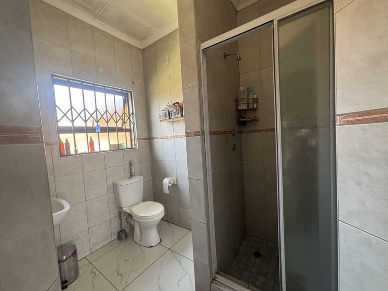 A functional bathroom featuring a shower enclosure, toilet, and wash basin with tiled finishes and a security gate on the window.
