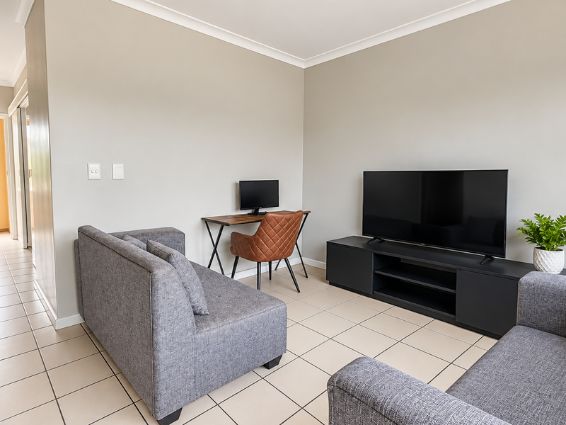 A well-maintained living room featuring neutral walls, tiled flooring, and access to a hallway leading to an outdoor balcony area.
