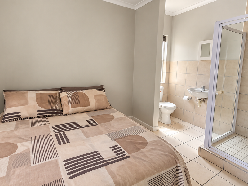 A bedroom featuring an open-plan en-suite bathroom with a shower enclosure, wall-mounted basin, and toilet, finished with neutral wall paint and ceramic tiling.