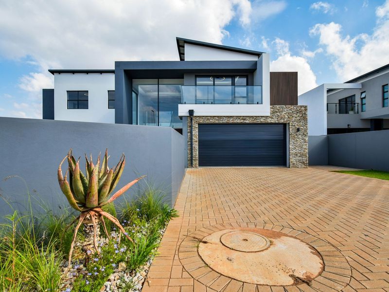 Modern double-story residential facade featuring a paved driveway, integrated double garage, stone cladding accents, and a glass-railed balcony.