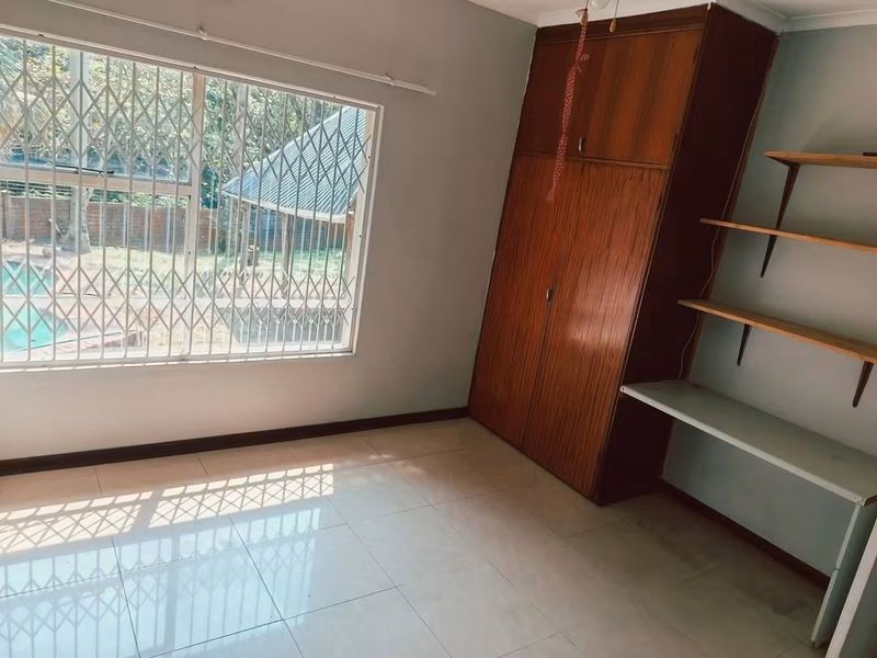 A bright, empty bedroom featuring tiled flooring, a built-in wooden wardrobe, and integrated shelving, with security bars on the window.