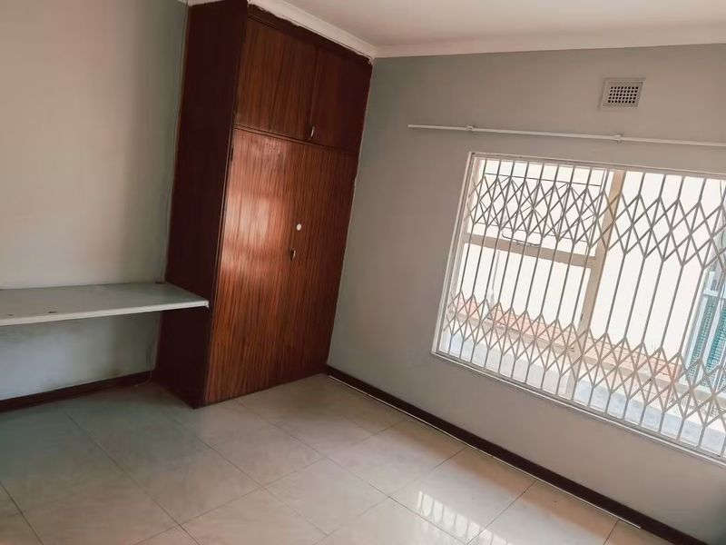 A bright, empty bedroom featuring tiled flooring, a built-in wooden wardrobe, a built-in desk, and a window fitted with a security gate.