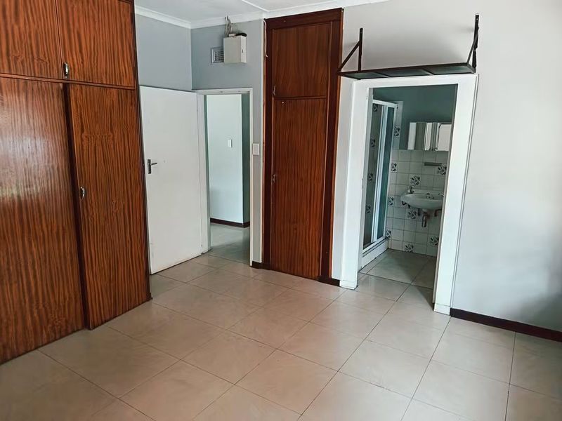 Empty bedroom featuring built-in wooden cupboards, tiled flooring, and an attached en-suite bathroom with a shower and pedestal basin.