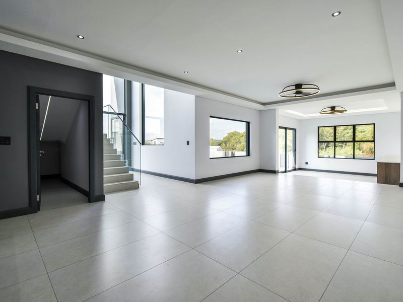 Spacious open-plan living area featuring large format floor tiles, modern recessed lighting, a feature staircase with glass balustrade, and expansive windows.