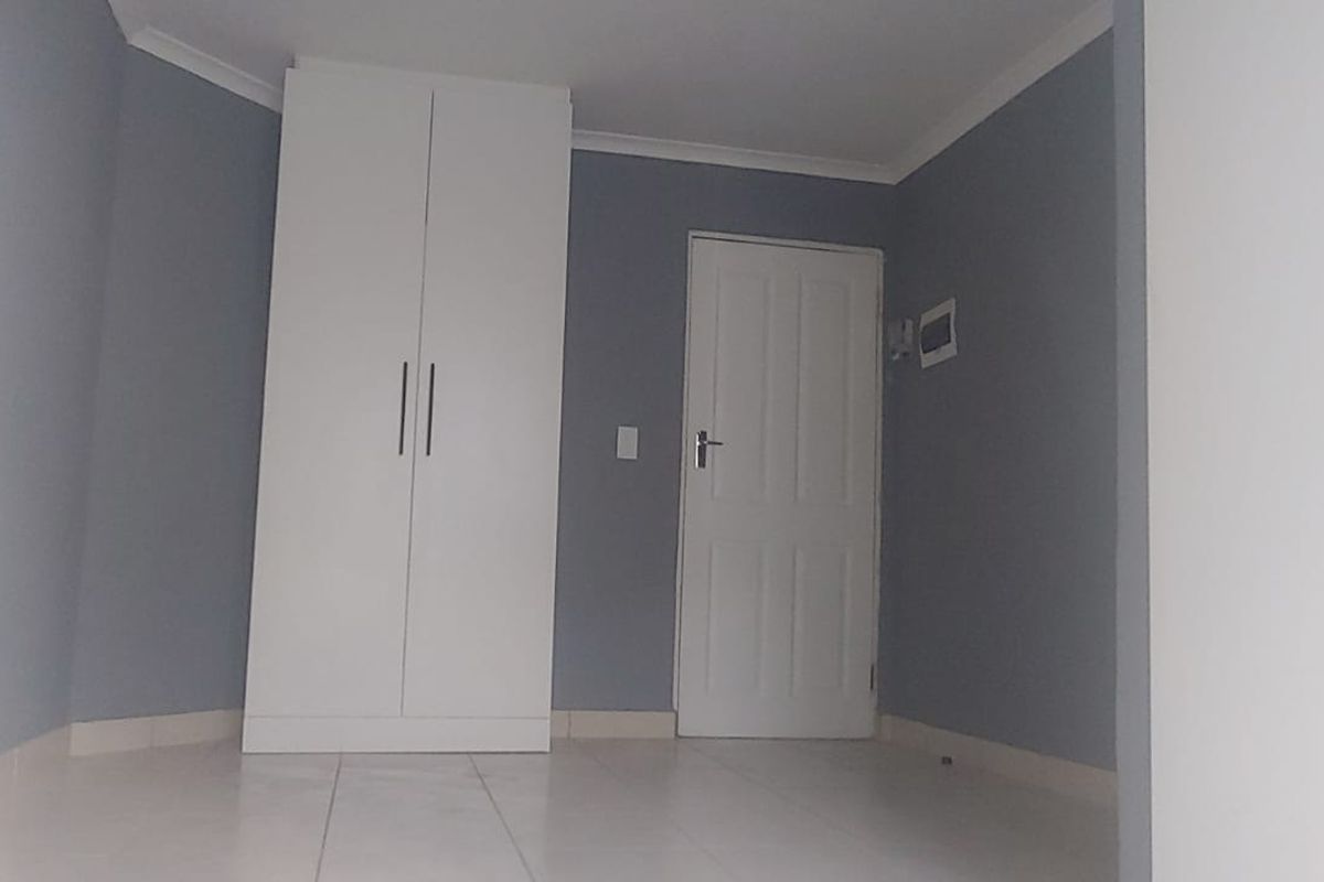 A clean, empty bedroom featuring neutral grey walls, white built-in wardrobe, tiled flooring, and a standard internal door.