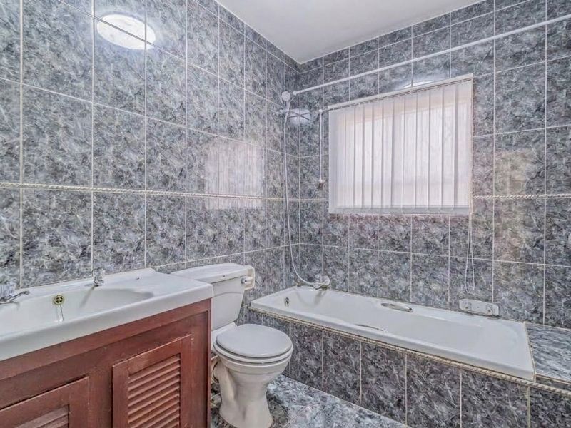 A functional bathroom featuring full-height marble-effect wall tiling, a built-in vanity, a standard bathtub with shower mixer, and a toilet.