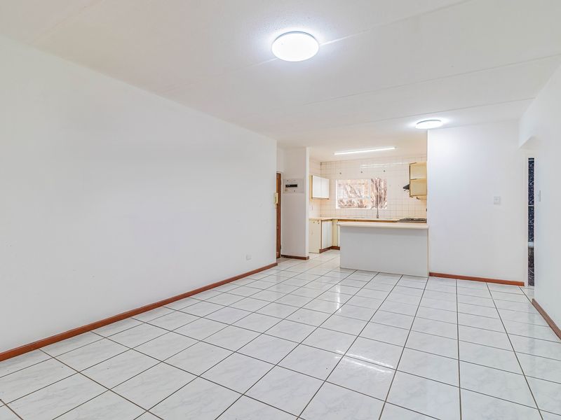 An open-plan living and kitchen area featuring white tiled flooring, neutral walls, and functional kitchen cabinetry with a breakfast counter.