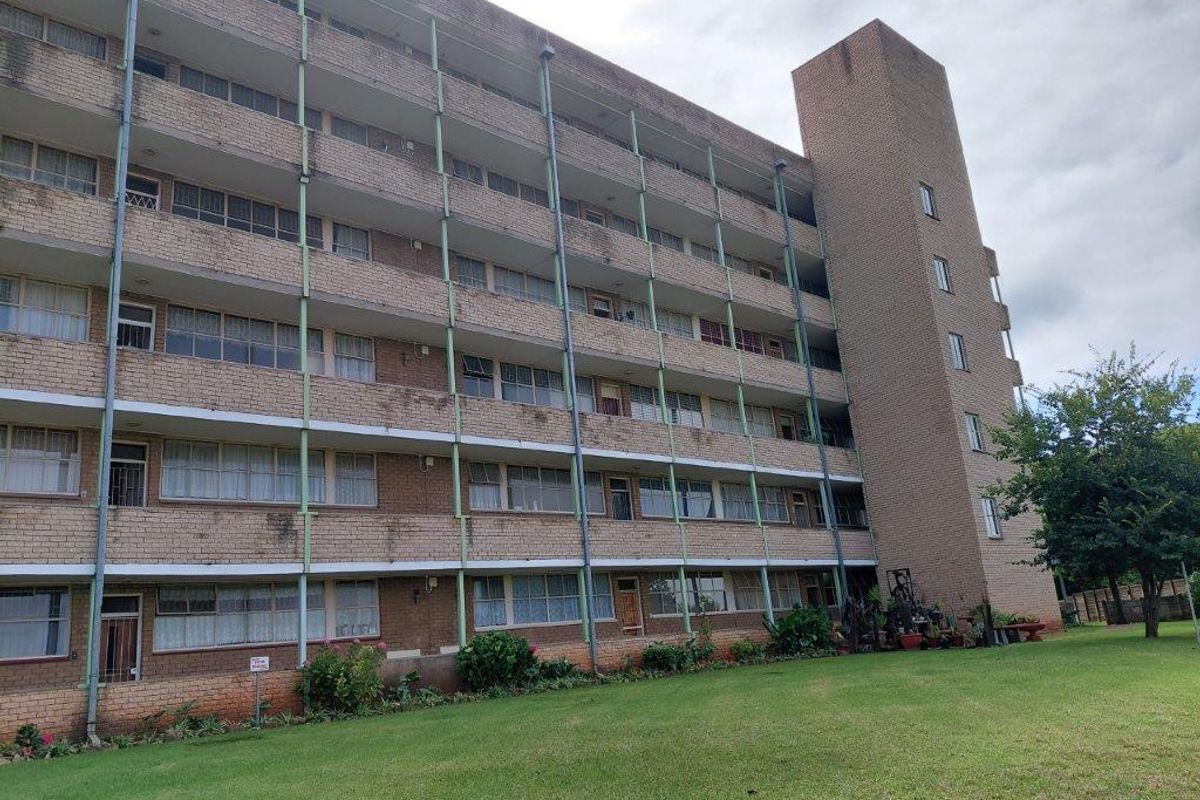 A multi-story brick apartment building featuring exterior balconies with metal supports, set against a well-maintained lawn area.