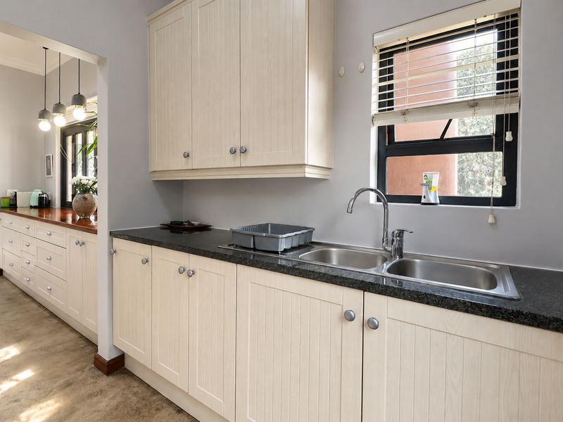 A well-maintained scullery featuring light wood-grain cabinetry, dark granite countertops, a double stainless steel sink, and polished concrete flooring.