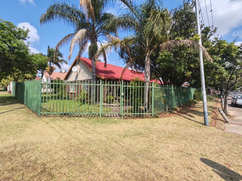 Front view of a brick house with a red tiled roof, enclosed by green metal fencing and surrounded by mature palm trees.