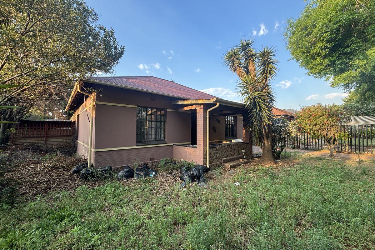 Rear view of a detached house featuring a corrugated metal roof, covered patio area, and surrounding garden space requiring landscaping maintenance.
