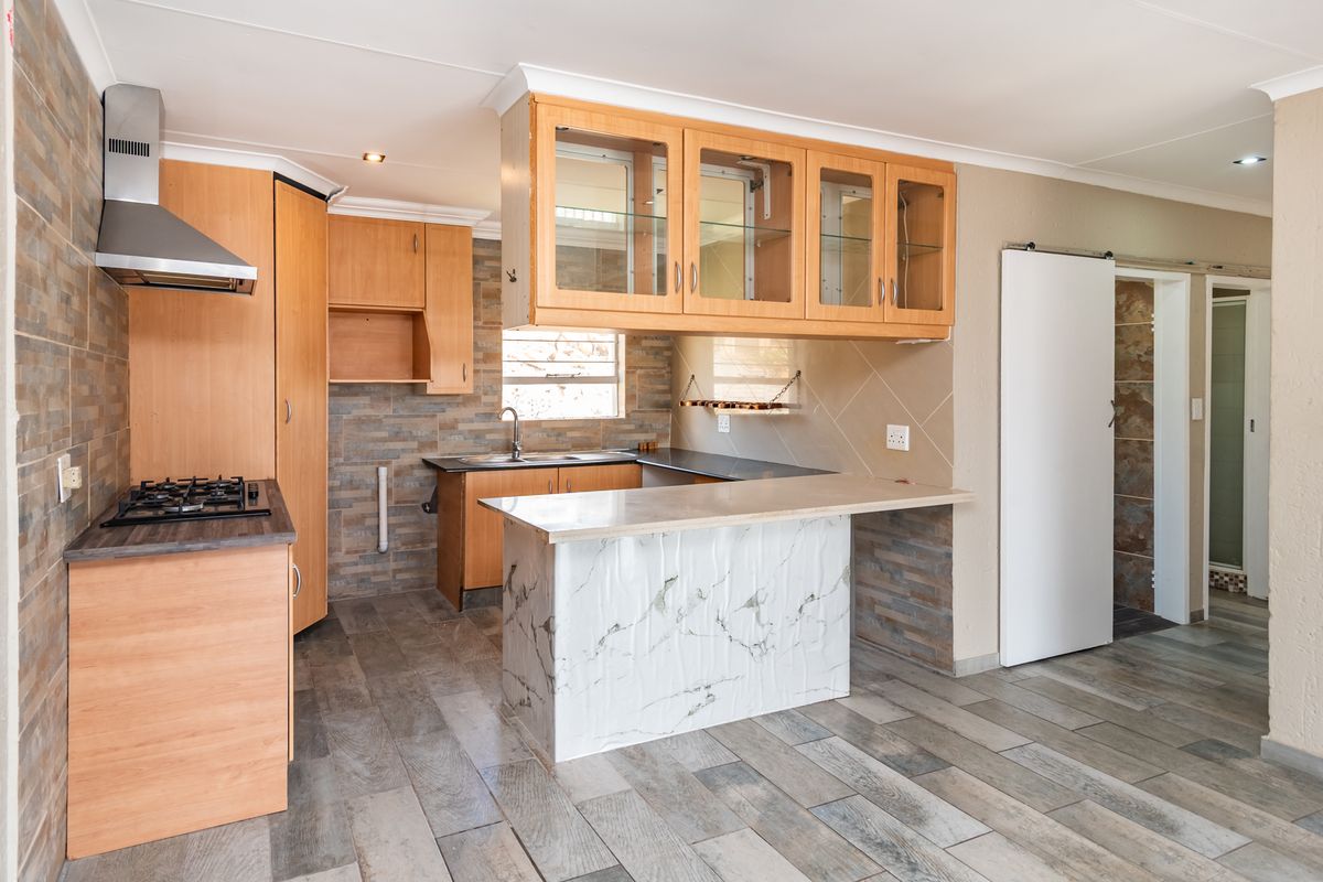 A functional kitchen featuring wood-finish cabinetry, a gas hob with extractor, stone-look wall cladding, and a marble-effect breakfast counter in an open-plan layout.