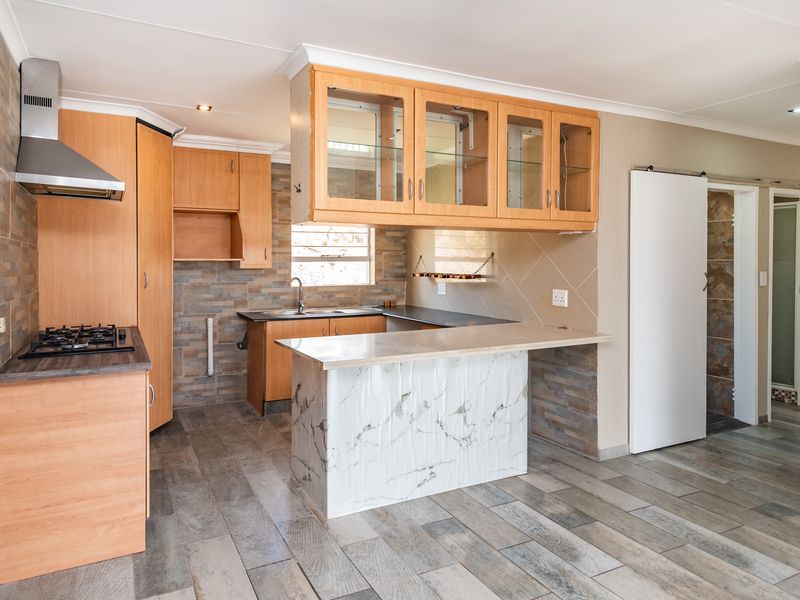 A functional kitchen featuring wood-finish cabinetry, a gas hob with extractor, stone-look wall cladding, and a marble-effect breakfast counter in an open-plan layout.
