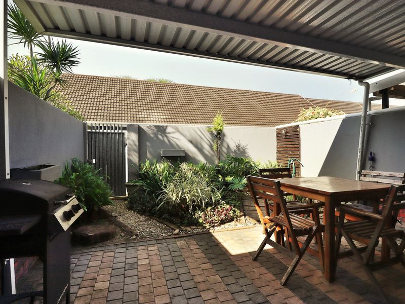 A covered outdoor patio area featuring paved flooring, a corrugated metal roof, and a private garden space with a pedestrian gate.