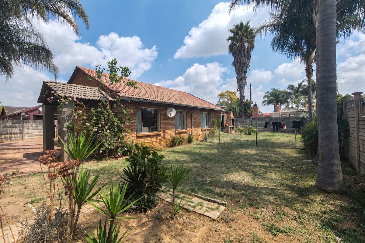 Rear view of a brick house featuring a tiled roof, a built-in outdoor braai, and a spacious garden with established trees and boundary walling.