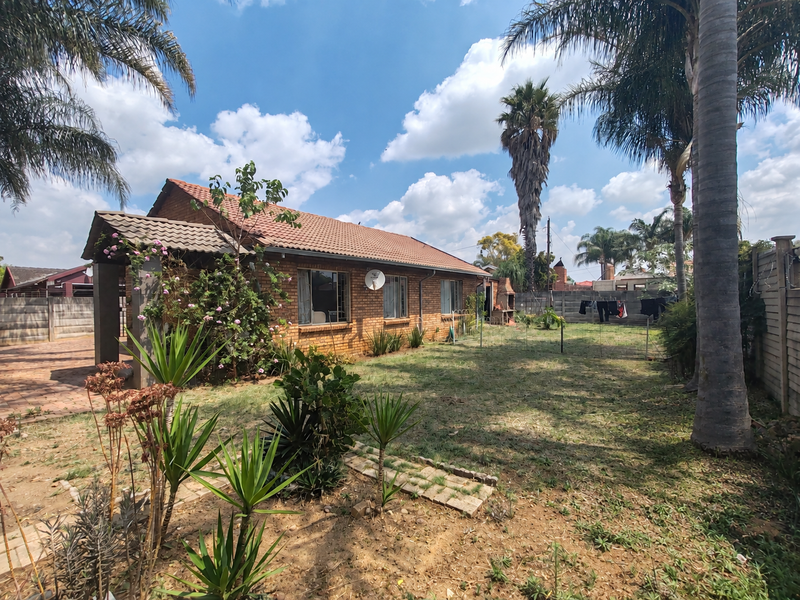 Rear view of a brick house featuring a tiled roof, a built-in outdoor braai, and a spacious garden with established trees and boundary walling.
