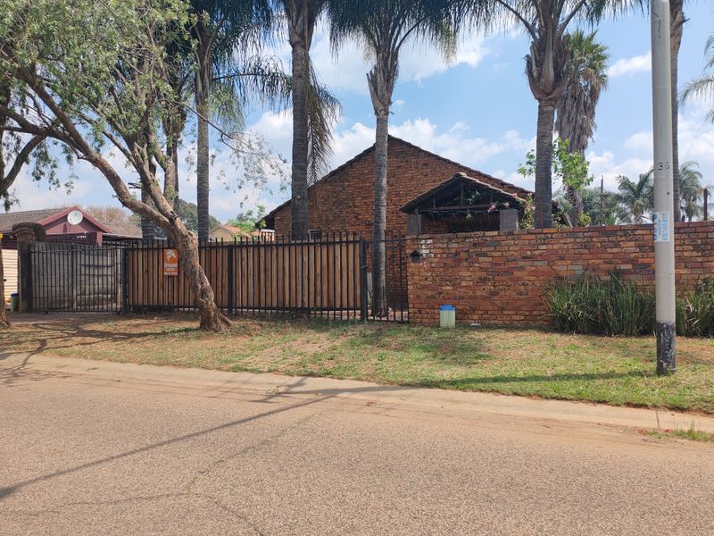 Street view of the property featuring a brick boundary wall, metal security fencing, and mature palm trees along the front perimeter.