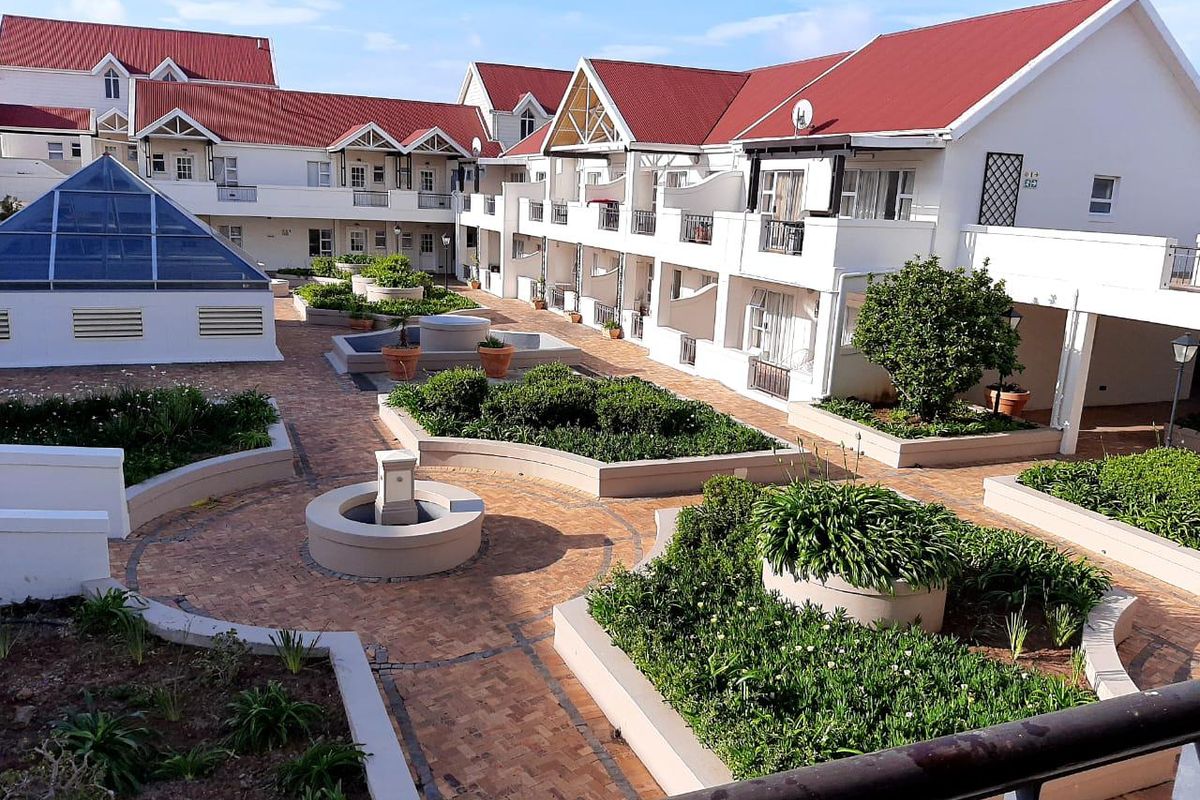 A well-maintained apartment complex courtyard featuring paved walkways, landscaped garden beds, water features, and multiple residential balconies under a pitched roof structure.