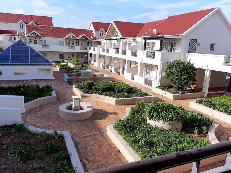 A well-maintained apartment complex courtyard featuring paved walkways, landscaped garden beds, water features, and multiple residential balconies under a pitched roof structure.