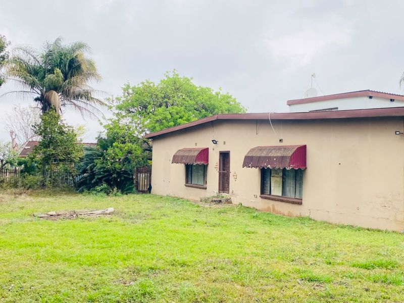 Rear view of the property featuring a large lawn area, weathered exterior walls, and metal window awnings.