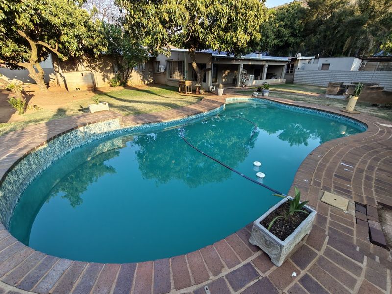 A backyard pool area featuring a curved swimming pool with brick coping, surrounded by a paved patio and a covered outdoor living space.