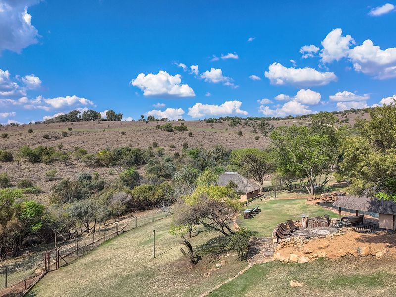 Aerial view of a rural property featuring thatched roof structures, a stone fire pit area, and expansive landscaped grounds set against a natural landscape.