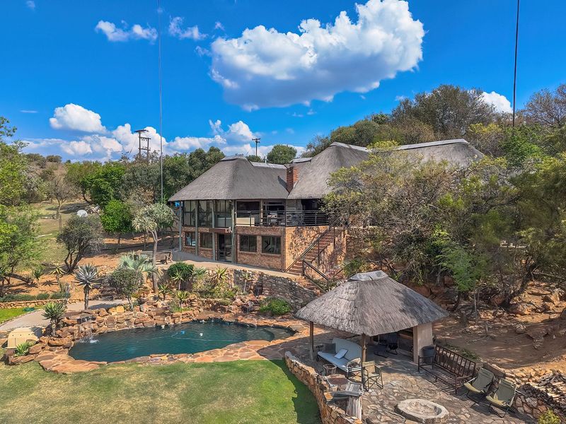 Aerial view of a multi-level residence featuring a thatched roof, stone-surrounded swimming pool, and a landscaped garden with a thatched gazebo.