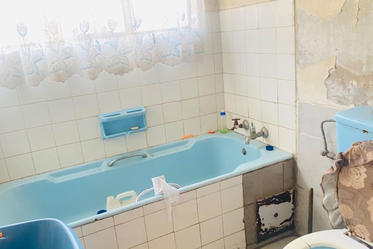 A dated bathroom featuring a blue bathtub and toilet, requiring significant renovation due to damaged wall finishes and worn tiling.